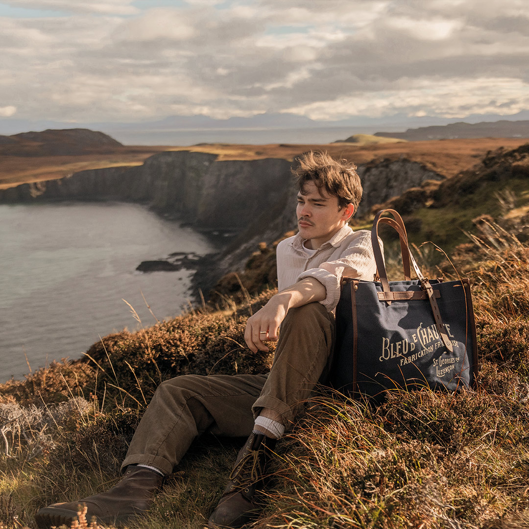 Un homme est allongé au bord des falaise écossaise, un cabas bleu est posé a coté de lui 