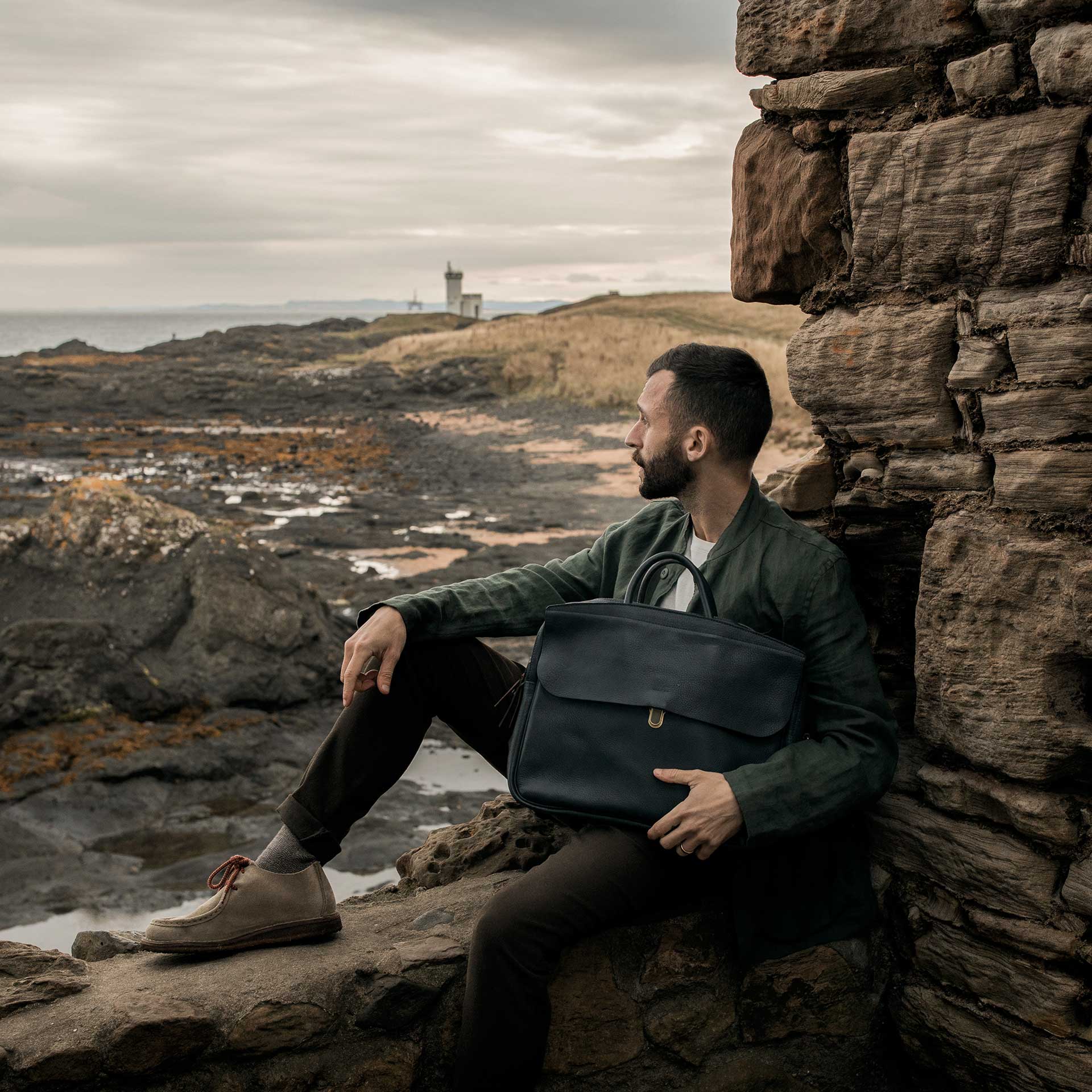 Un homme est assis sur des ruines et regarde le paysage devant lui, il porte un sac en cuir bleu